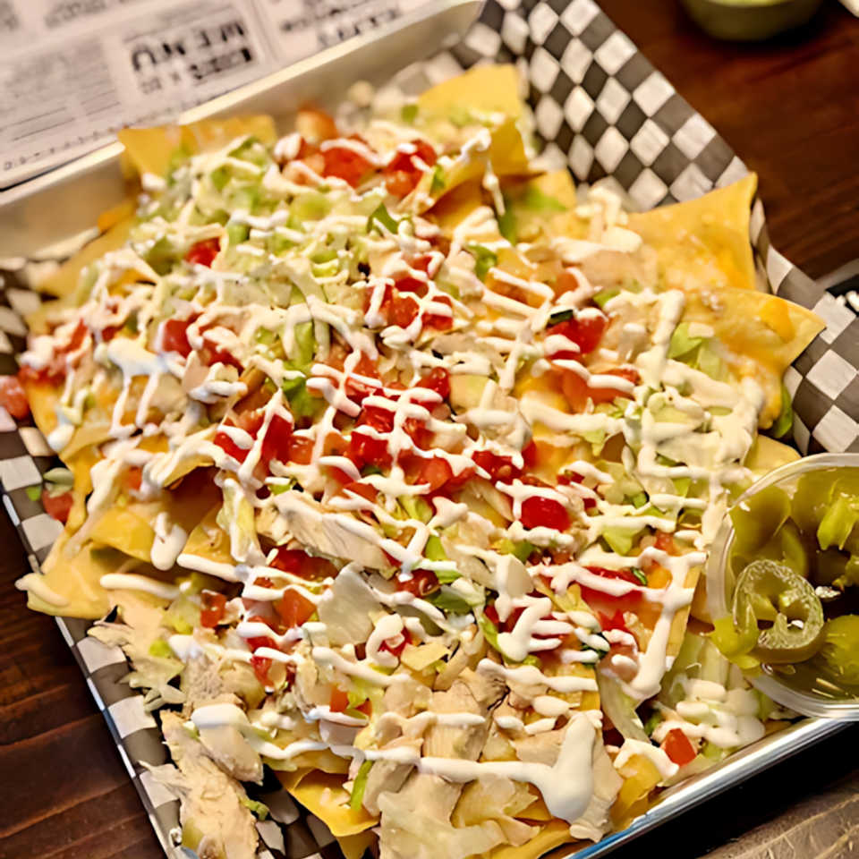 Loaded nachos topped with chicken, lettuce, tomatoes, cheese, sour cream, guacamole, and jalapeños on a checkered-lined tray.