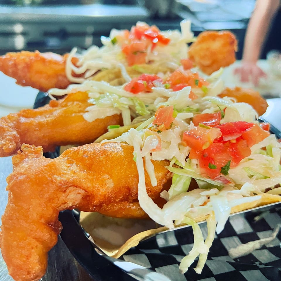 Crispy golden fish tacos topped with shredded lettuce and diced tomatoes, served in a black basket lined with checkered paper.