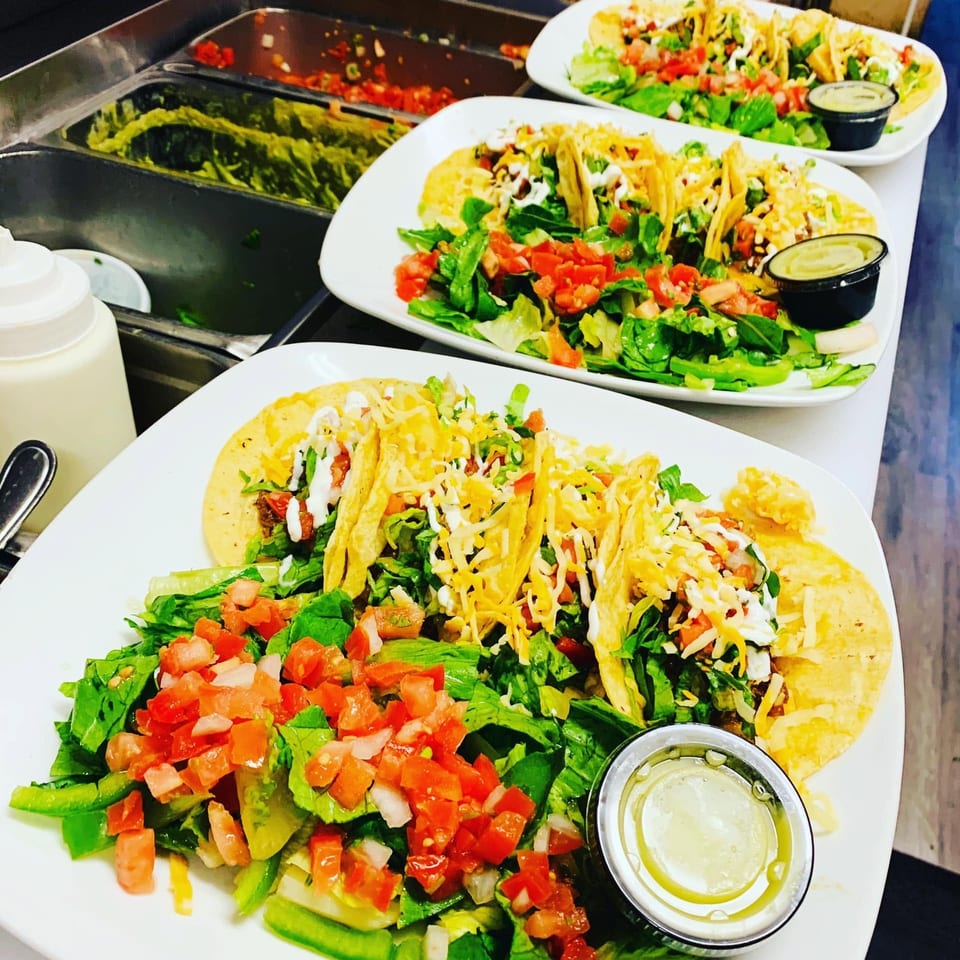 Plates of crunchy tacos topped with lettuce, tomatoes, cheese, and sour cream alongside fresh salads and dipping sauces.