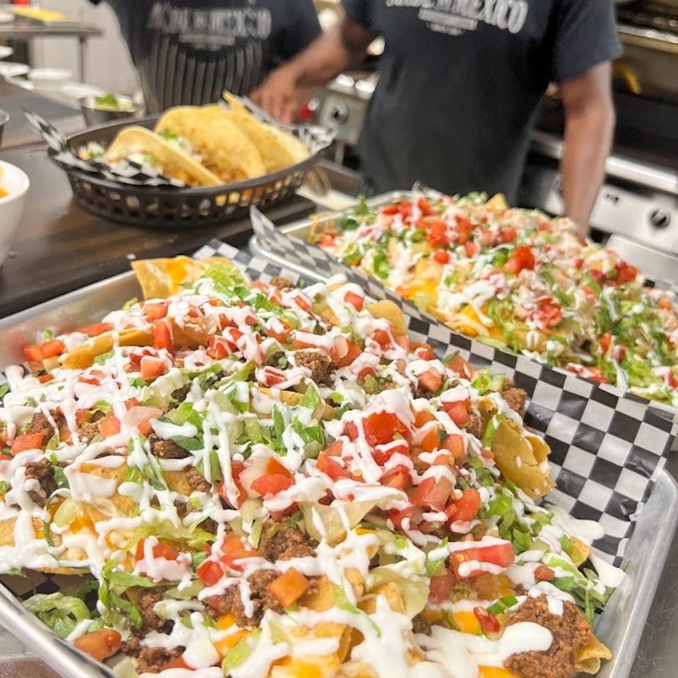 Loaded nachos topped with seasoned beef, lettuce, tomatoes, cheese, and creamy sauce, with tacos and restaurant staff in the kitchen.