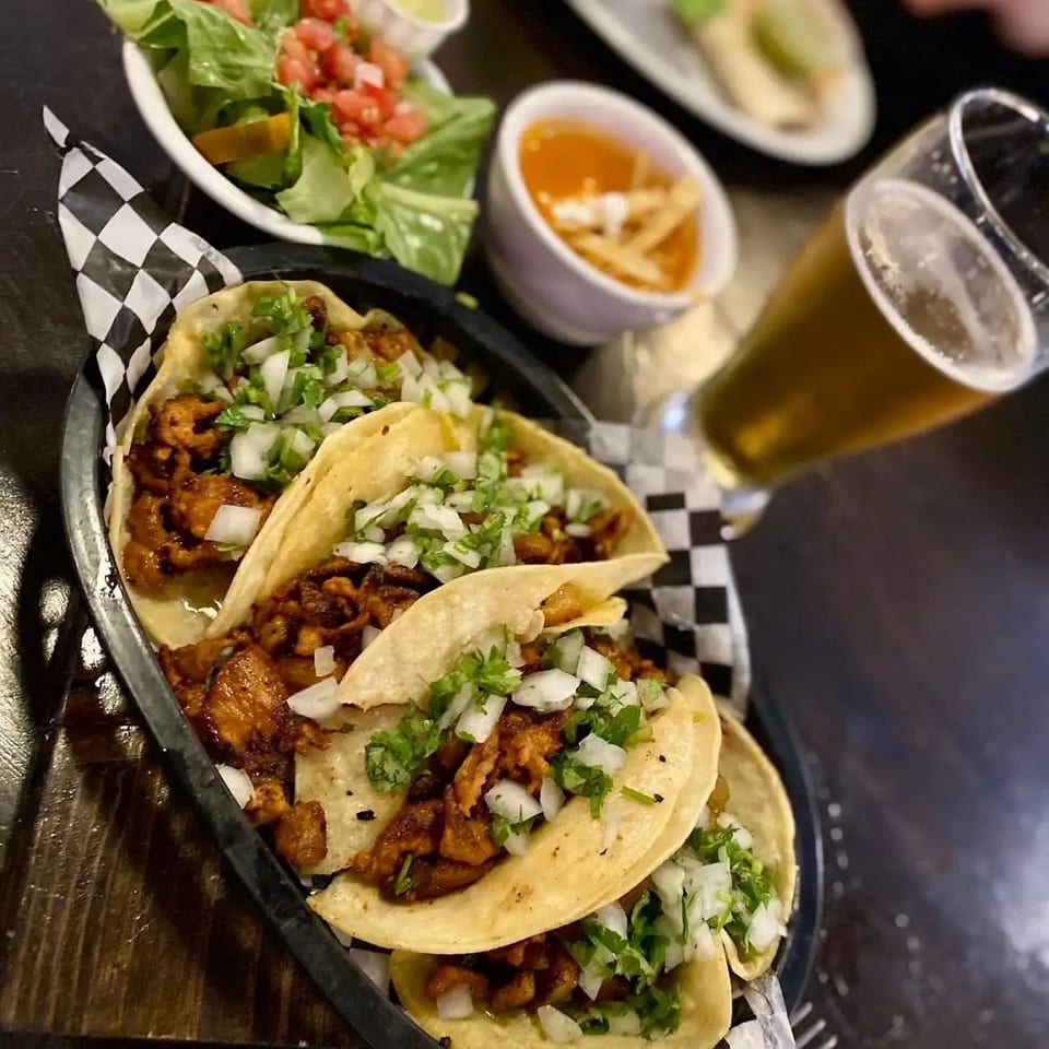 Four soft tacos filled with seasoned meat, onions, and cilantro served with salad, salsa, and a glass of beer.