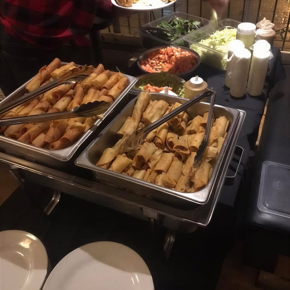 Buffet station with trays of golden spring rolls, fresh vegetables, sauces, serving utensils, and stacked plates.
