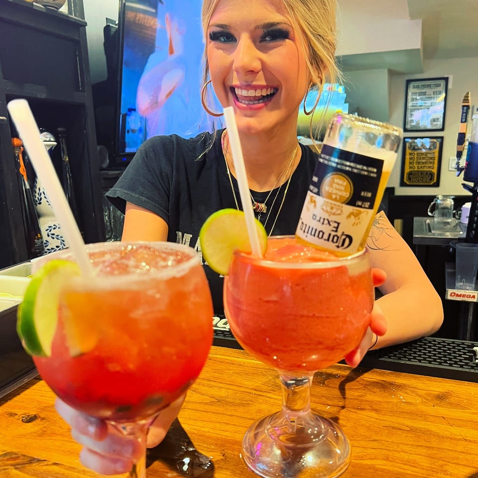 Smiling bartender presents two red frozen cocktails garnished with lime, one topped with an inverted Corona Extra bottle.