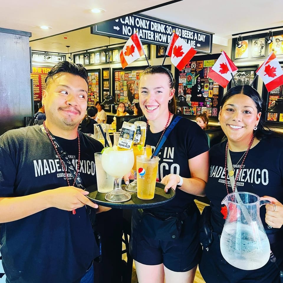 Three smiling restaurant staff pose with Canadian flags, drinks, and a water pitcher inside a lively, memorabilia-covered bar.