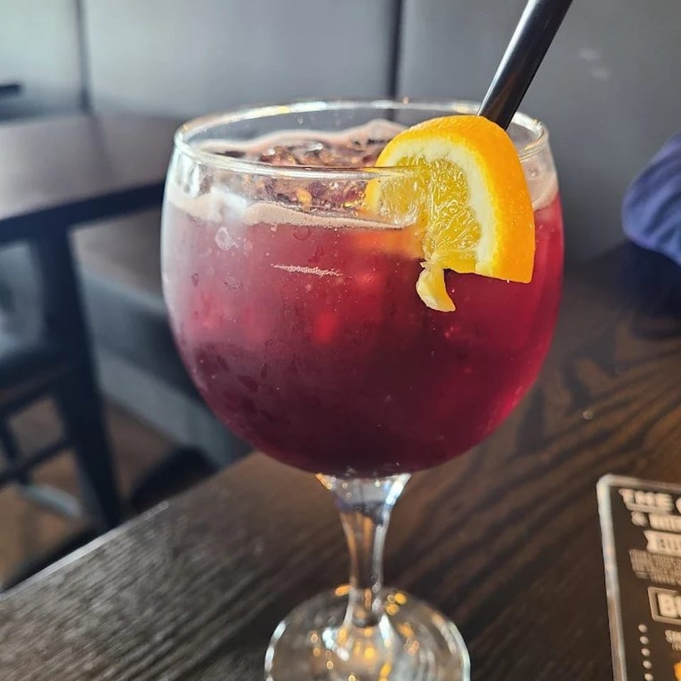 Deep red fruit cocktail in a stemmed glass, garnished with an orange slice and served with a black straw at a restaurant table.
