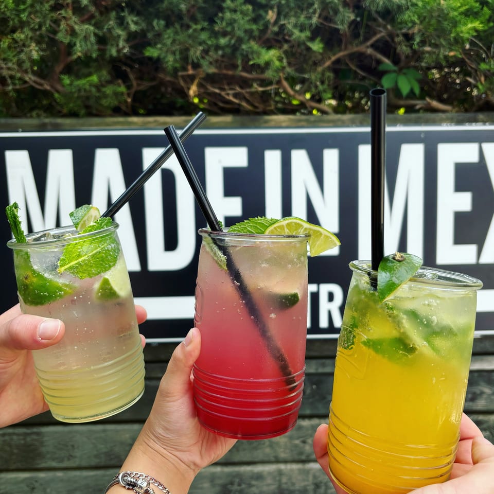 Three colorful lime-and-mint cocktails in clear glasses are held outdoors against a restaurant sign and greenery.