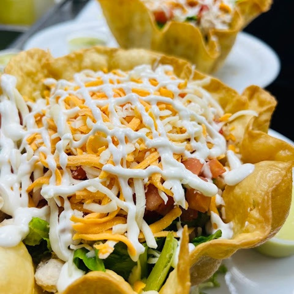Crispy tortilla bowl filled with salad, vegetables, shredded cheese, diced tomatoes, and creamy white dressing, with another topped bowl behind it.