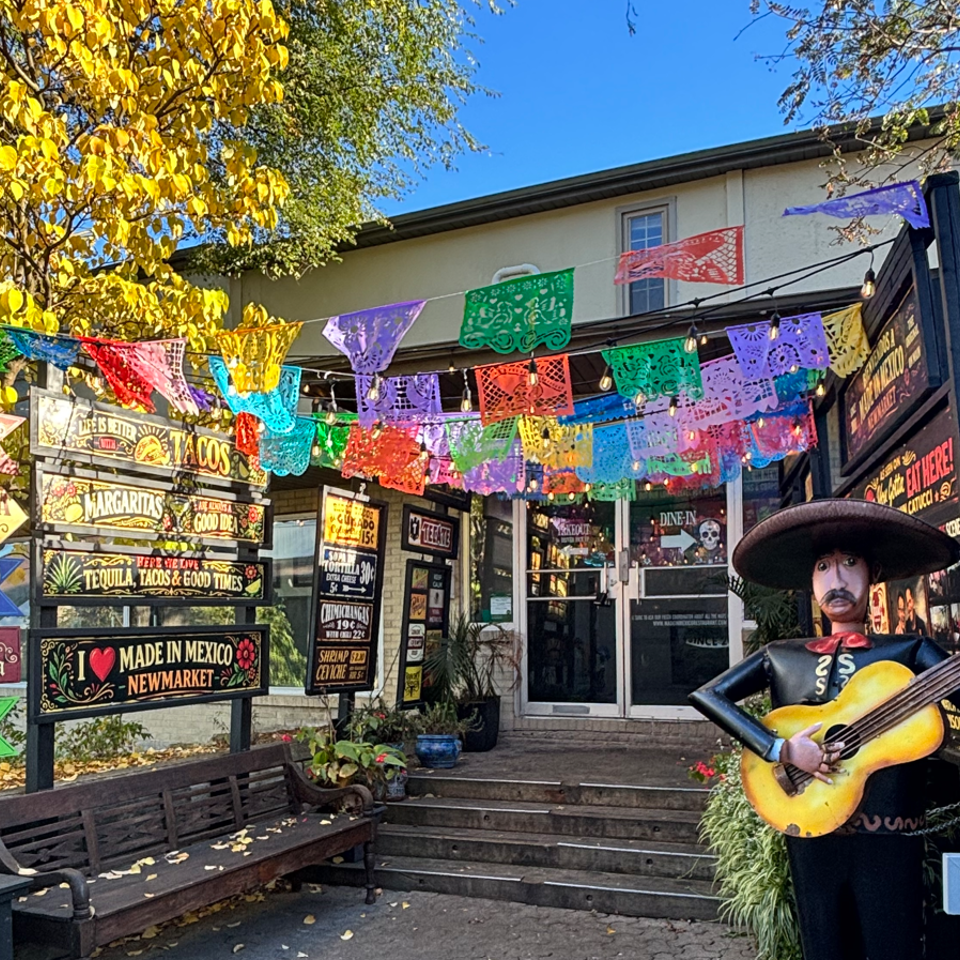 Colorful Mexican restaurant entrance decorated with papel picado banners, regional signs, string lights, and a mariachi statue playing guitar.