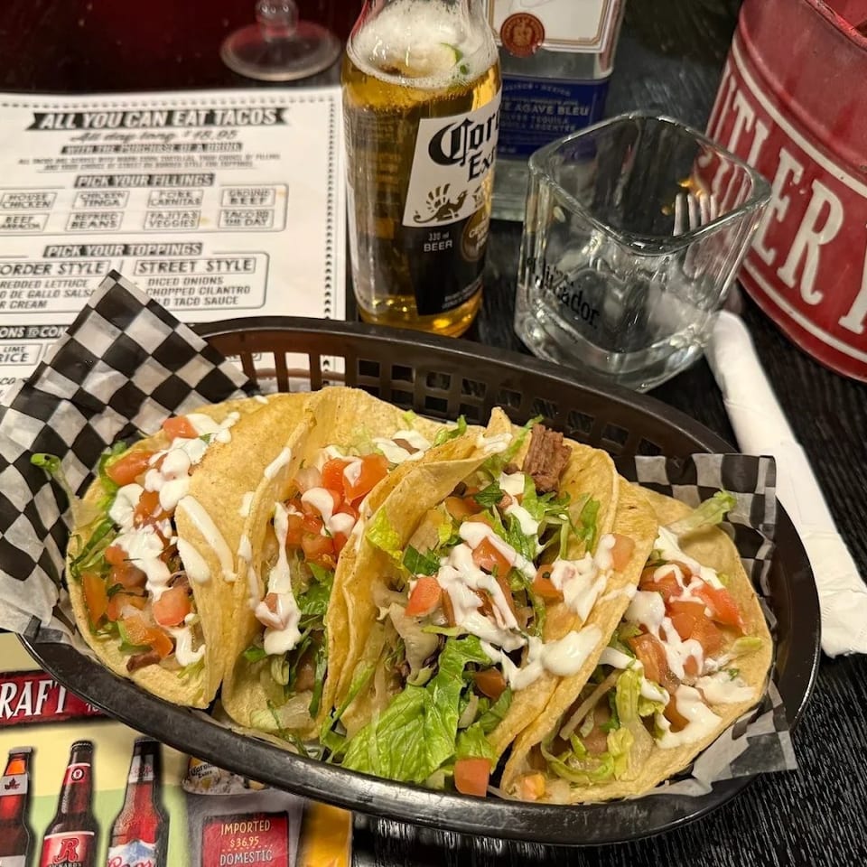 Basket of crispy tacos topped with lettuce, diced tomatoes, white sauce, and meat beside a bottle of Corona beer.